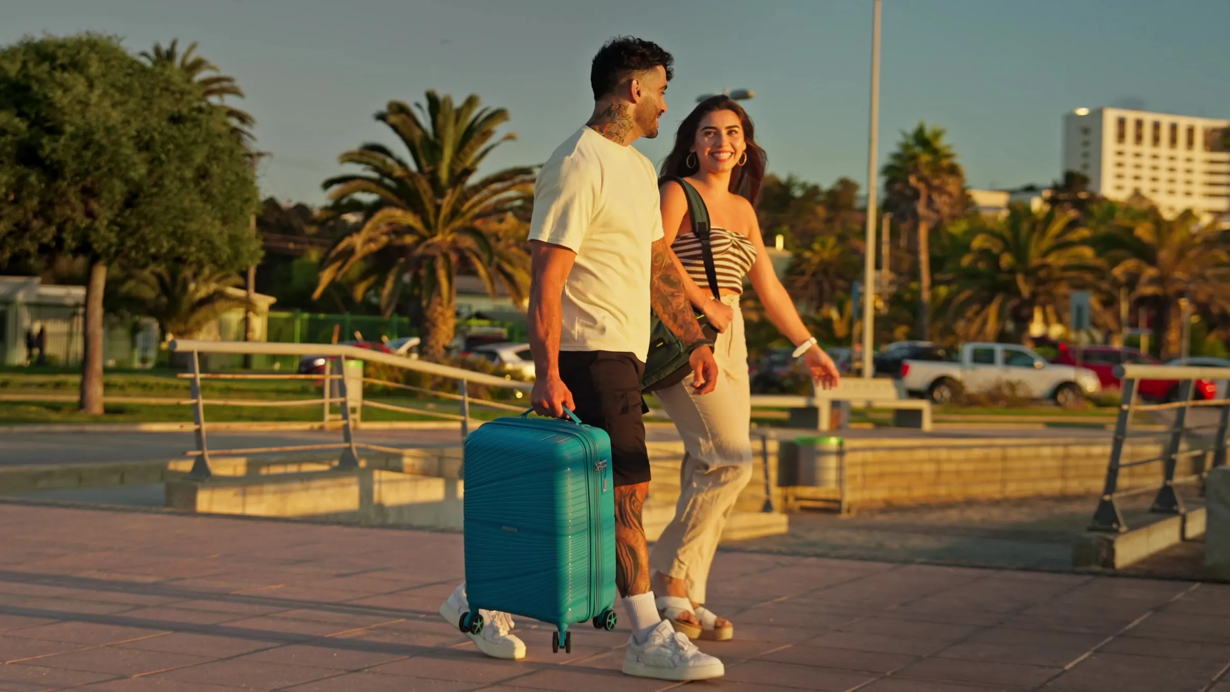 Toma de la campaña de verano de American Tourister: una pareja camina con una maleta turquesa por la costanera
